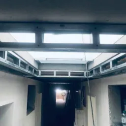 Metalowa konstrukcja sufitu podwieszanego w trakcie montażu, widoczne profile i elementy mocujące, surowy stan ścian, w tle widok na kolejne pomieszczenie.