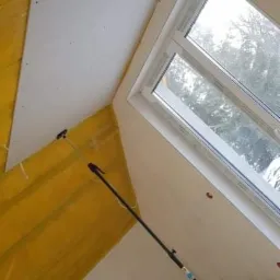 Montaż płyty gipsowo-kartonowej na poddaszu, widoczne fragmenty ocieplenia, okno dachowe i teleskopowa podpórka.