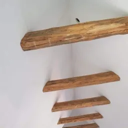 Poddasze z widocznymi drewnianymi belkami stropowymi, pomalowane na biało, z widocznym otworem w suficie i fragmentem drabiny w dolnym rogu.