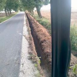 Wykopany rów wzdłuż drogi asfaltowej, widok z kabiny maszyny budowlanej, brązowa ziemia, drzewa w tle.