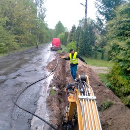 AMG Stefania wąsik - Pracownik w żółtej kamizelce niesie ciasto nad wykopem, w tle czerwony bus i drugi pracownik przy drodze, mokry asfalt i żółta koparka.