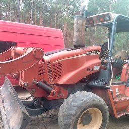 AMG Stefania wąsik - Pomarańczowa koparka Ditch Witch RT115 z lemieszem i wysokim kominem, zaparkowana obok czerwonego vana na tle lasu sosnowego.