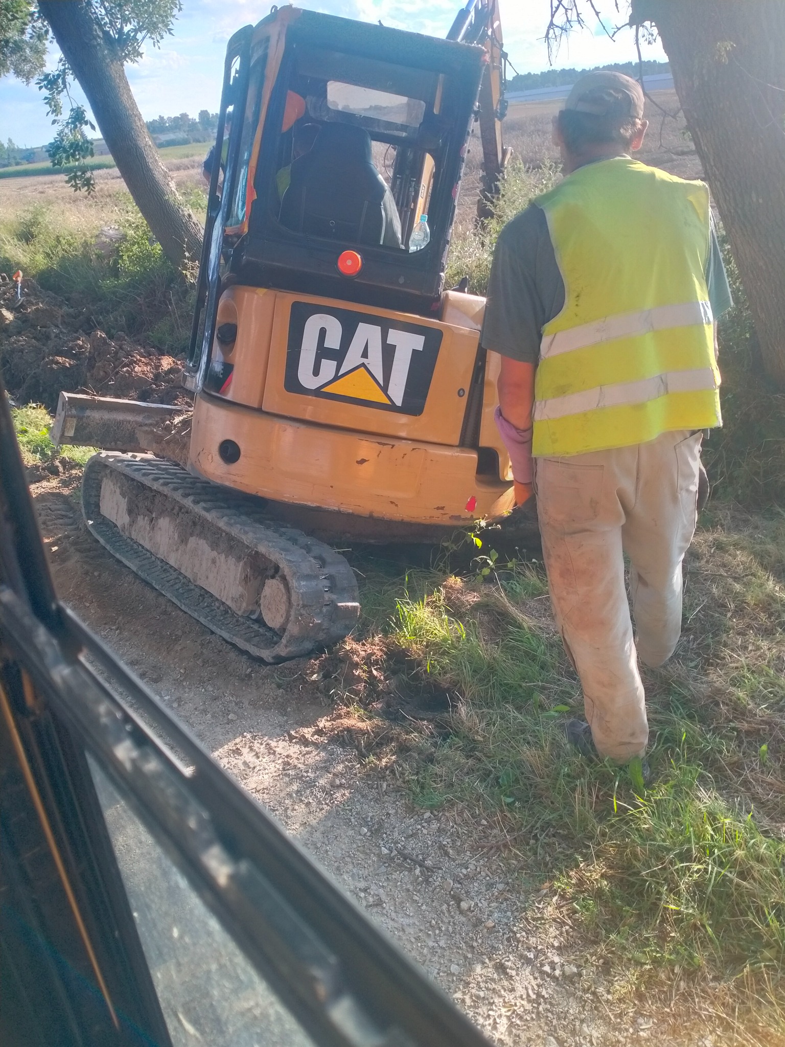 Operator w kamizelce odblaskowej stoi obok małej koparki CAT w trakcie prac ziemnych przy drzewach na polu.