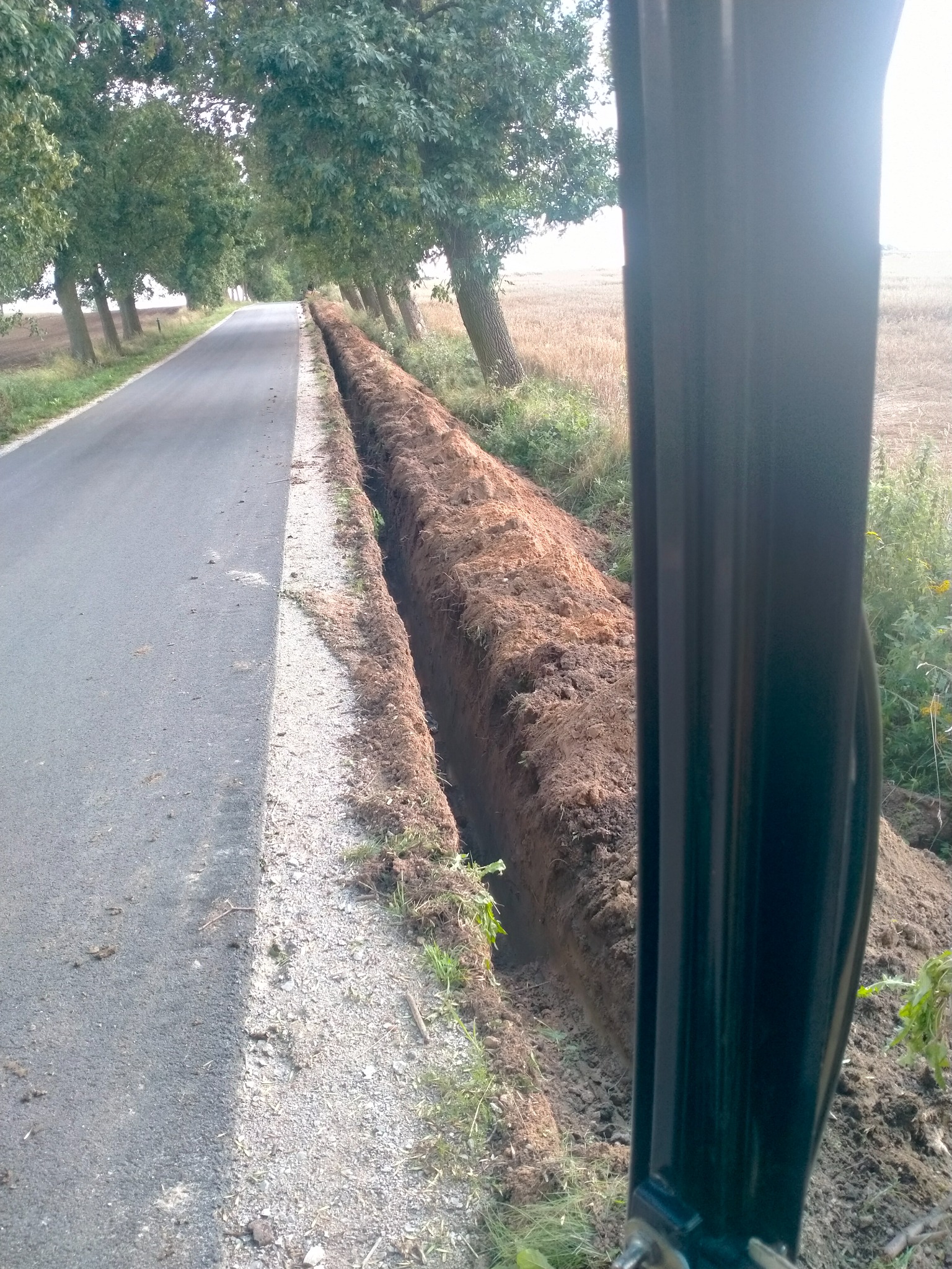 Wykopany rów wzdłuż drogi asfaltowej, widok z kabiny maszyny budowlanej, brązowa ziemia, drzewa w tle.