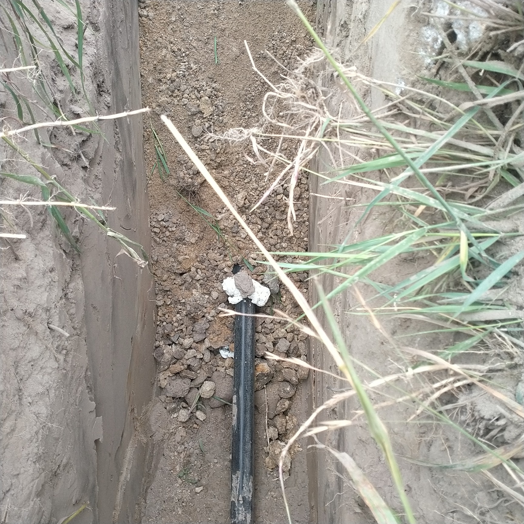 Rura telekomunikacyjna w wykopie, zabezpieczona pianką montażową, widok z góry na ziemię i trawę.