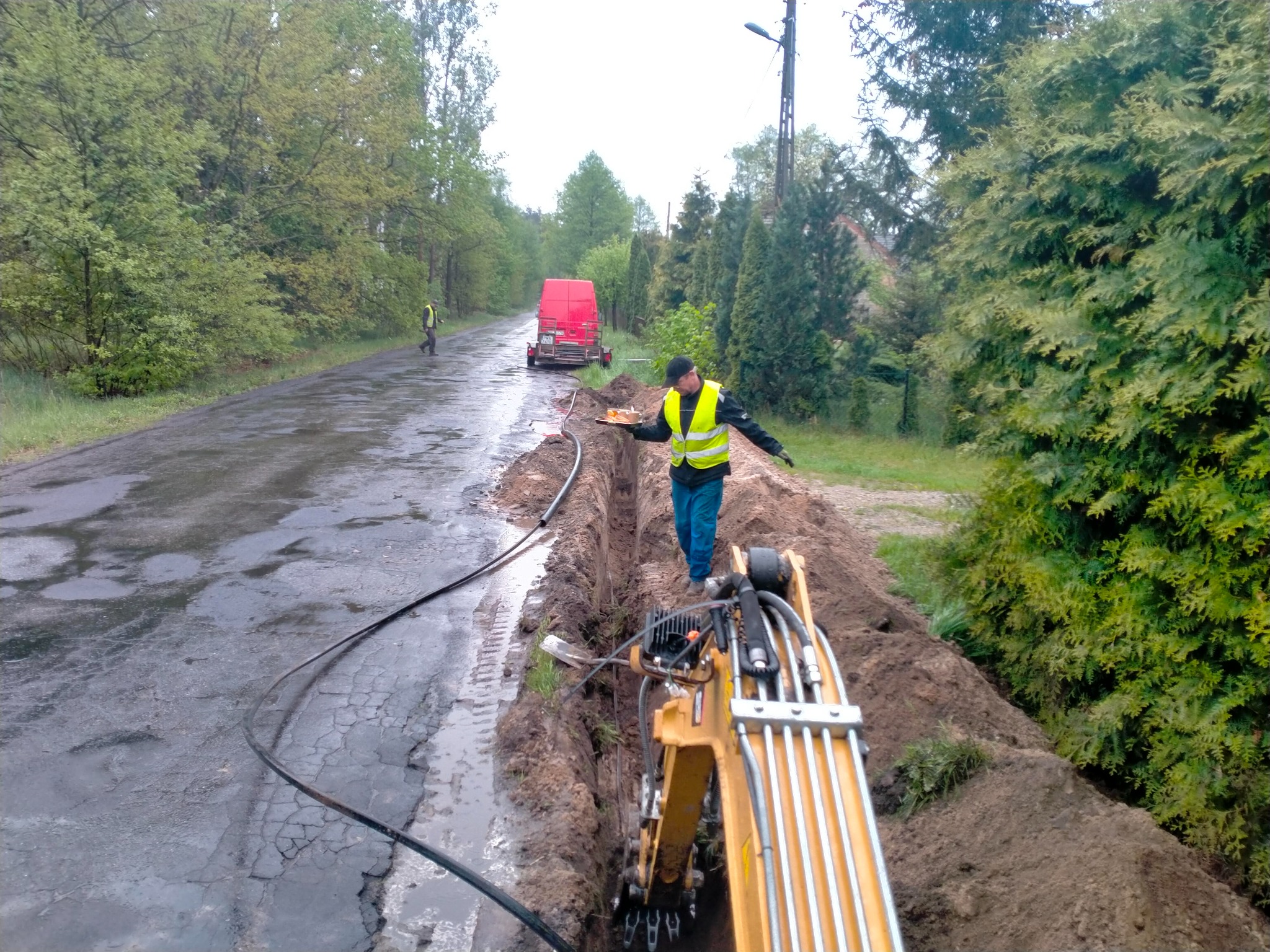 Pracownik w żółtej kamizelce niesie ciasto nad wykopem, w tle czerwony bus i drugi pracownik przy drodze, mokry asfalt i żółta koparka.