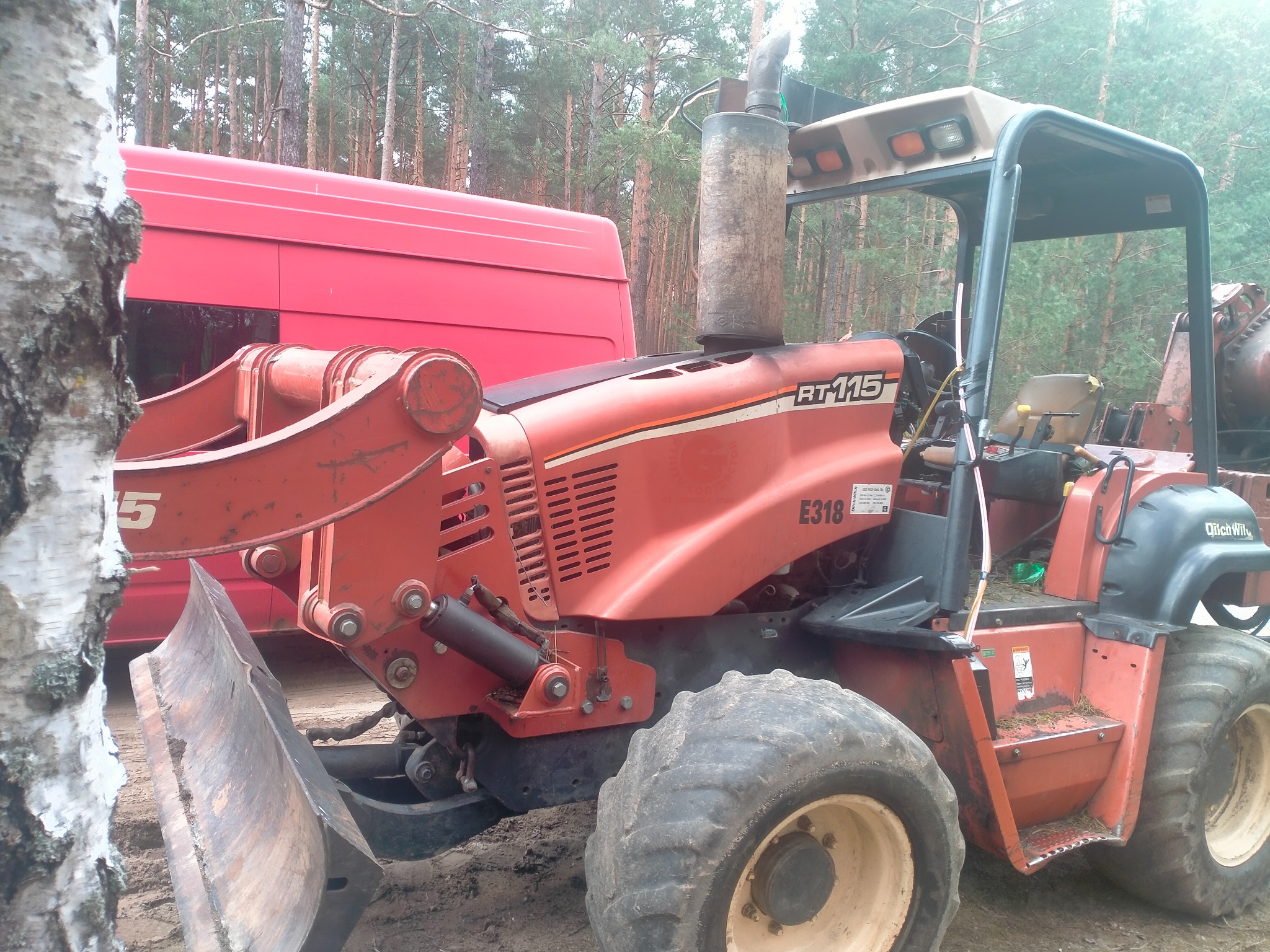 Pomarańczowa koparka Ditch Witch RT115 z lemieszem i wysokim kominem, zaparkowana obok czerwonego vana na tle lasu sosnowego.