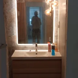 Wykończona łazienka z umywalką w szafce z jasnego drewna i podświetlanym lustrem, na ścianach płytki imitujące beton, widoczne niedociągnięcia w wykończeniu ściany po prawej stronie lustra.