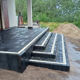 Nowoczesny taras z ciemnej kostki brukowej z jasnymi obrzeżami, prowadzący schodami w dół; w tle widoczny dom z kolumną i stos piasku.