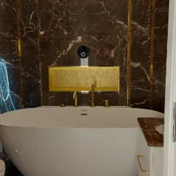 Luksusowa łazienka z wolnostojącą wanną, armaturą w kolorze złota i marmurowymi płytkami, oświetlona kryształowym żyrandolem.