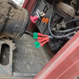 Otwarta komora silnika pojazdu z widocznym akumulatorem, bezpiecznikami i przewodami elektrycznymi, wszystko pokryte warstwą kurzu i brudu.