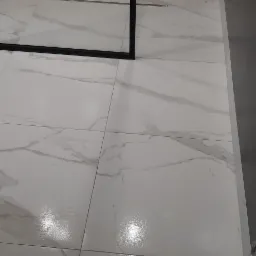 Białe, błyszczące płytki podłogowe imitujące marmur, ułożone w łazience z widoczną czarną ramą kabiny prysznicowej i dekoracyjną ścianą z drewnopodobnych paneli.