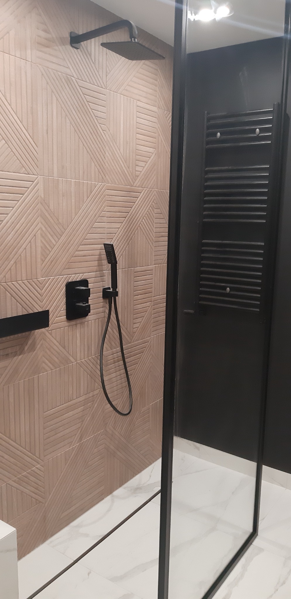 Nowoczesna kabina prysznicowa z czarnymi elementami, deszczownicą i słuchawką, wykończona płytkami o geometrycznym wzorze i grzejnikiem drabinkowym.