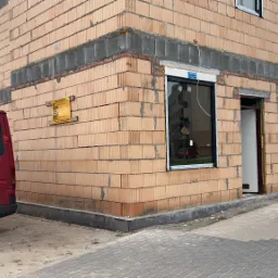 Niedokończony budynek z czerwonej cegły ceramicznej z zamontowanymi oknami i otwartymi drzwiami, widoczny czerwony van z tablicą rejestracyjną na tle.