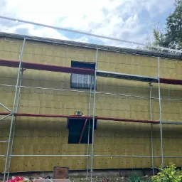 Ściana budynku w trakcie ocieplania wełną mineralną, widoczne rusztowanie i okna, fragment ogrodu w tle.