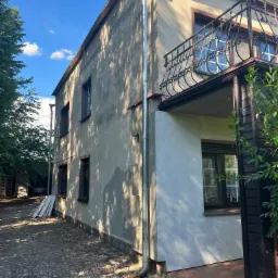 Dwupiętrowy dom z szarą elewacją przed ociepleniem, fragment ściany po remoncie z nowym tynkiem, widoczne rynny i balkon z ozdobną, metalową balustradą, kostka brukowa na podwórku.