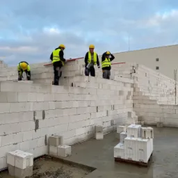 Budowa ścian z betonu komórkowego: murarze w żółtych kaskach i odblaskowych kamizelkach układają bloczki, widoczny zbrojony filar i palety z bloczkami na mokrej posadzce.