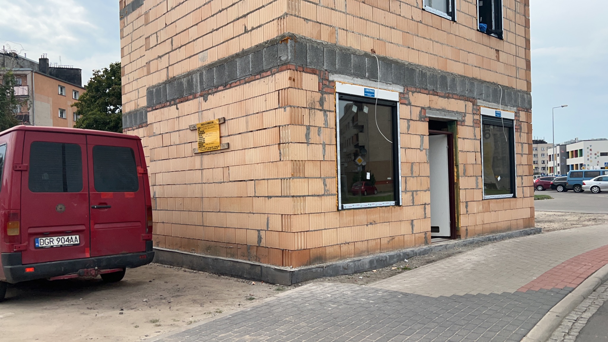Niedokończony budynek z czerwonej cegły ceramicznej z zamontowanymi oknami i otwartymi drzwiami, widoczny czerwony van z tablicą rejestracyjną na tle.