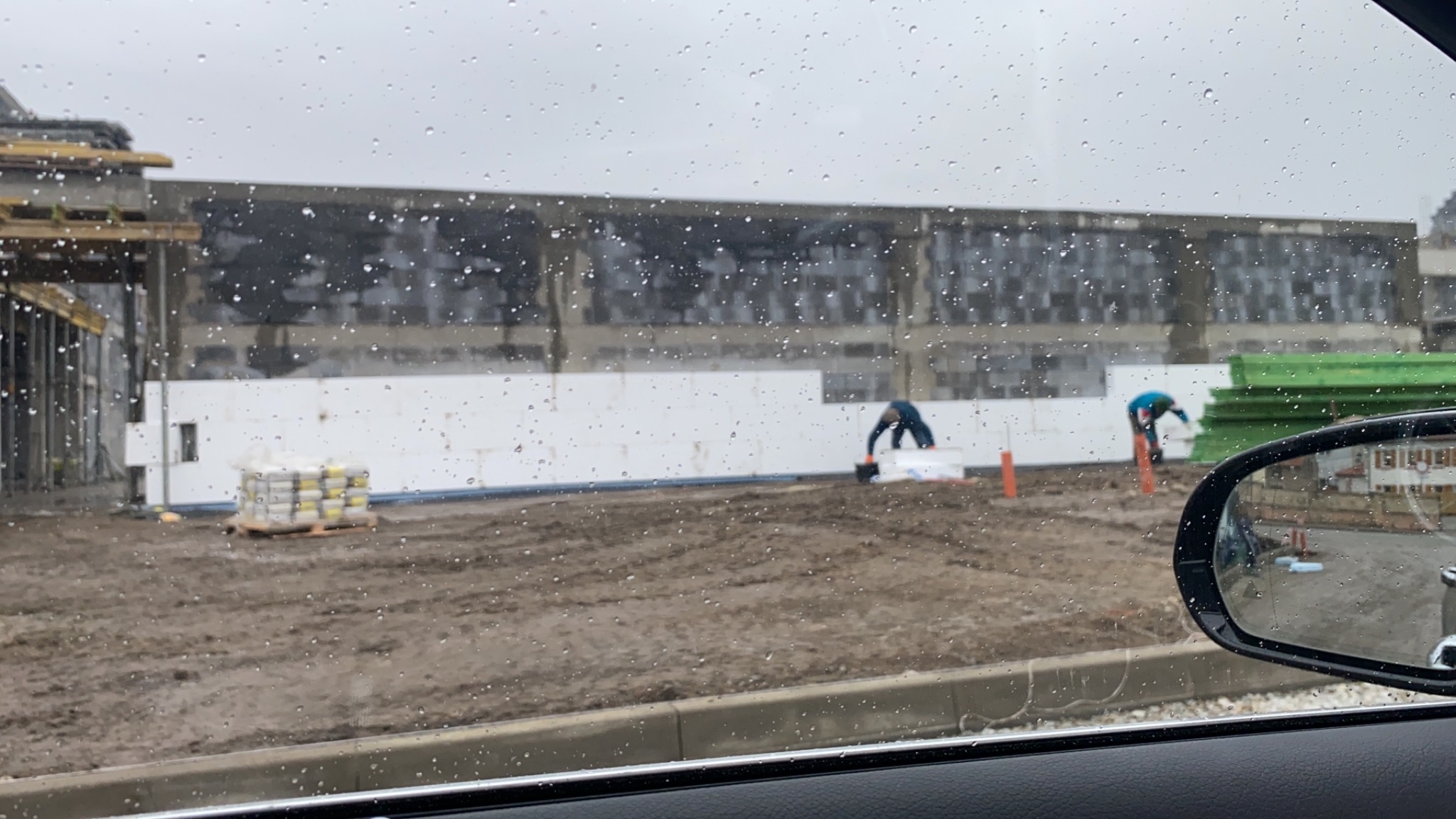 Praca na budowie w deszczowy dzień: dwóch pracowników montuje białe płyty izolacyjne na ścianie budynku, widok przez mokrą szybę samochodu.