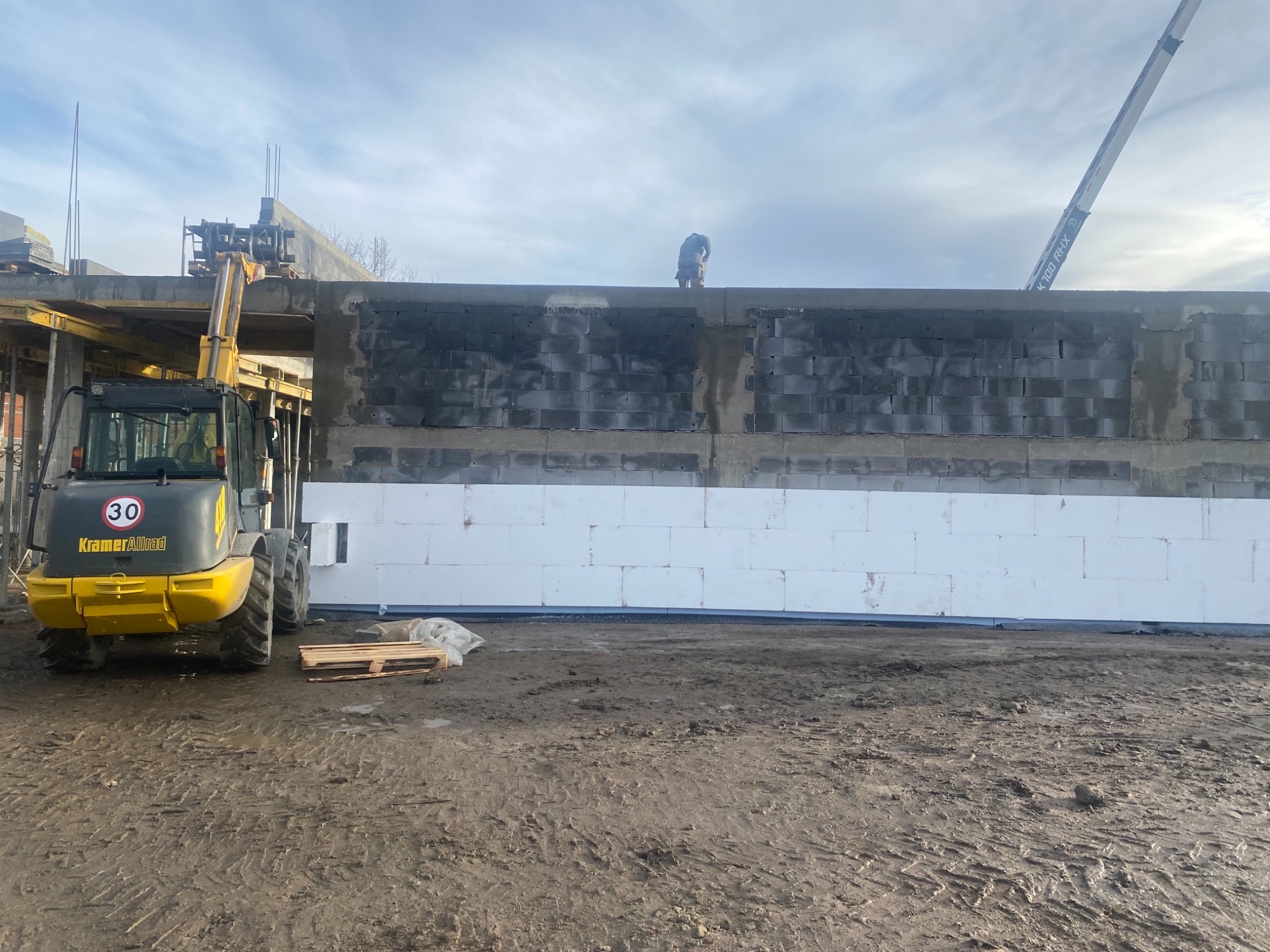 Budowa domu w trakcie: ściany z szarego bloczka częściowo ocieplone białym styropianem, widoczny operator na górze ściany, żółta ładowarka KramerAllrad po lewej stronie, teren budowy błotnisty.