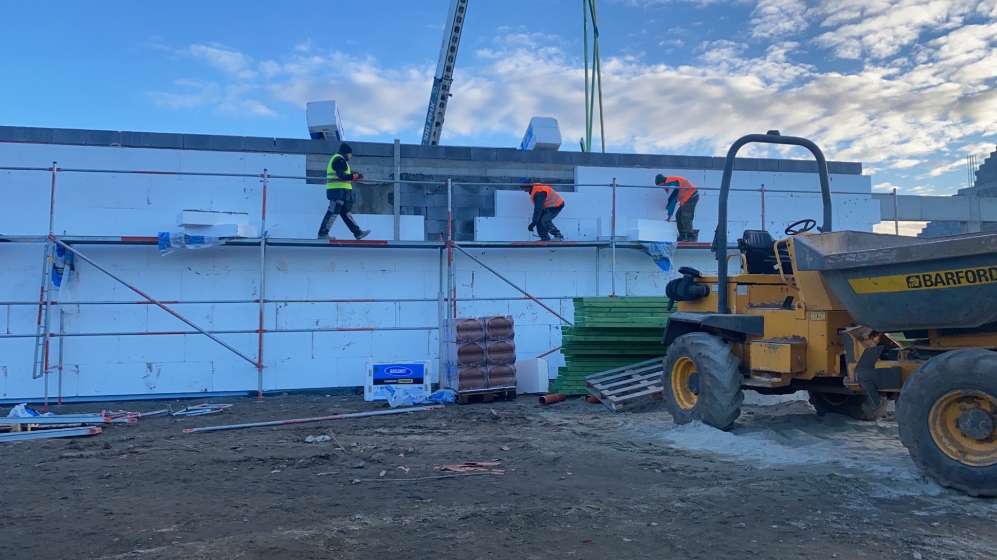 Pracownicy na rusztowaniu montują białe płyty izolacyjne na ścianie budowanego domu. W tle widoczny dźwig i żółta wywrotka.
