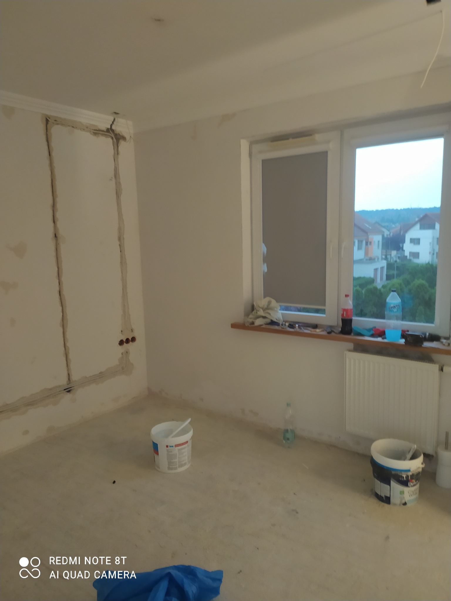 Przygotowane do malowania wnętrze pokoju z widocznymi śladami po instalacjach elektrycznych, dwa wiadra z farbą na podłodze, okno z widokiem na zabudowę mieszkalną.