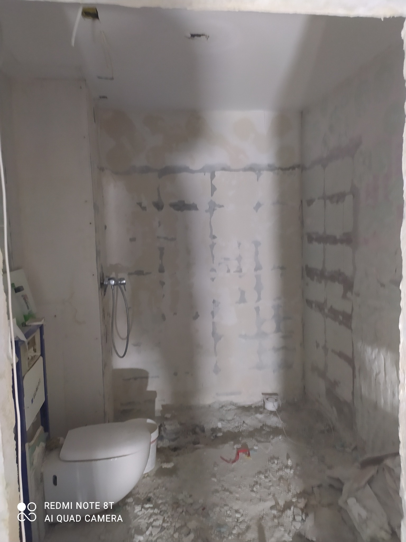 Łazienka w trakcie remontu: surowe ściany po skuciu płytek, widoczne ślady szpachlowania, zamontowana podtynkowa instalacja WC i bateria prysznicowa, gruz na podłodze, przygotowanie do dalszych...