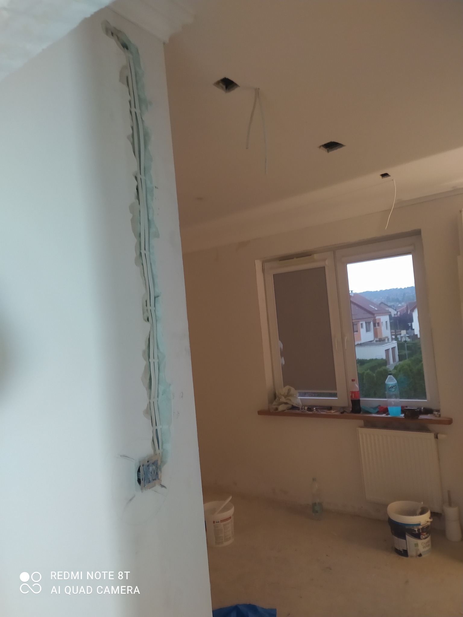 Wnętrze w trakcie remontu: pionowy bruzda na ścianie wypełniona gipsem, odsłonięte kable elektryczne w suficie, widok z okna na zabudowę jednorodzinną, wiaderka z farbą na podłodze.