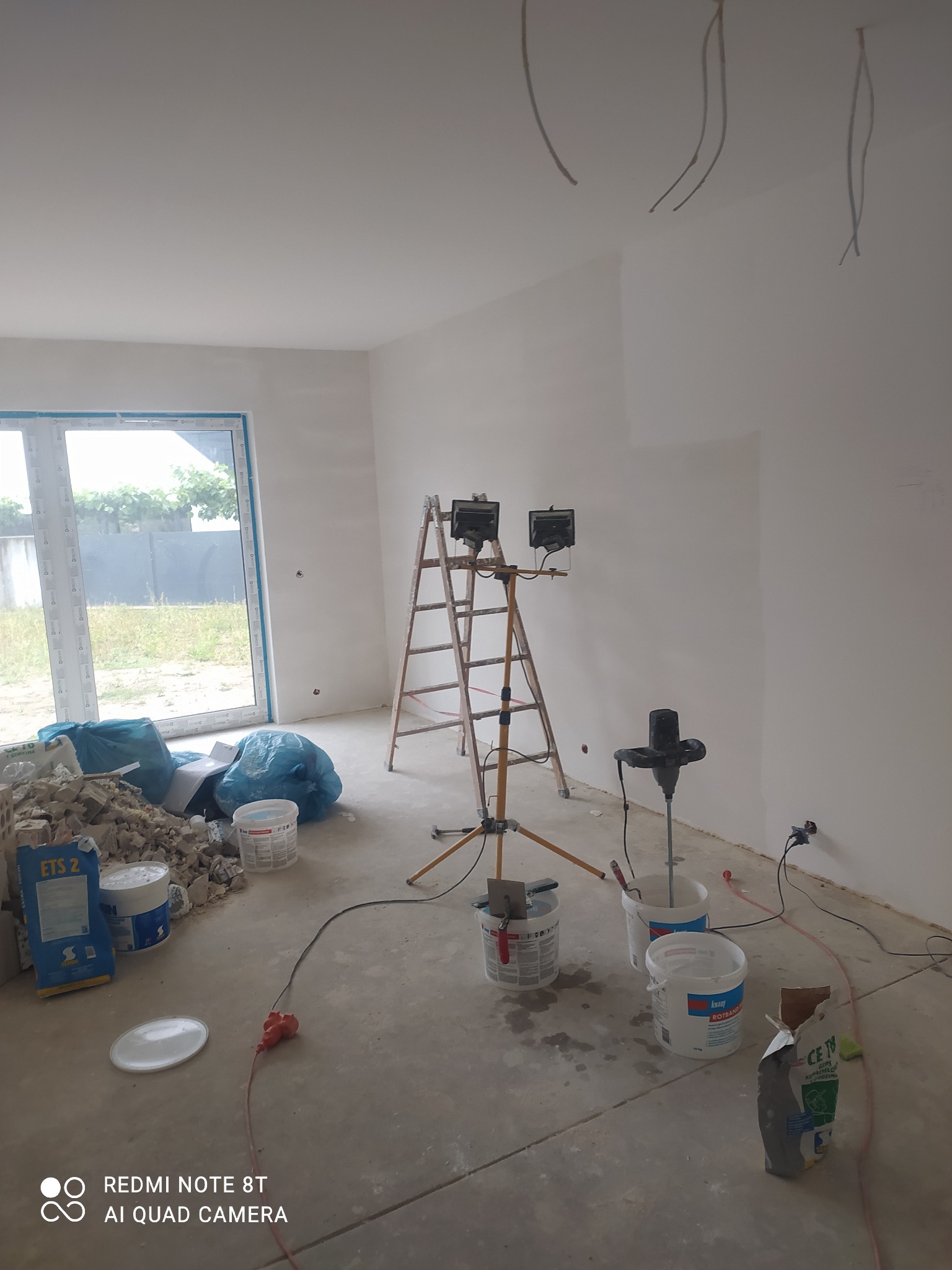 Wnętrze w trakcie prac wykończeniowych: ściany przygotowane do malowania, widoczne narzędzia, drabina z oświetleniem, worki i wiadra z materiałami budowlanymi, oraz niezabezpieczona instalacja...