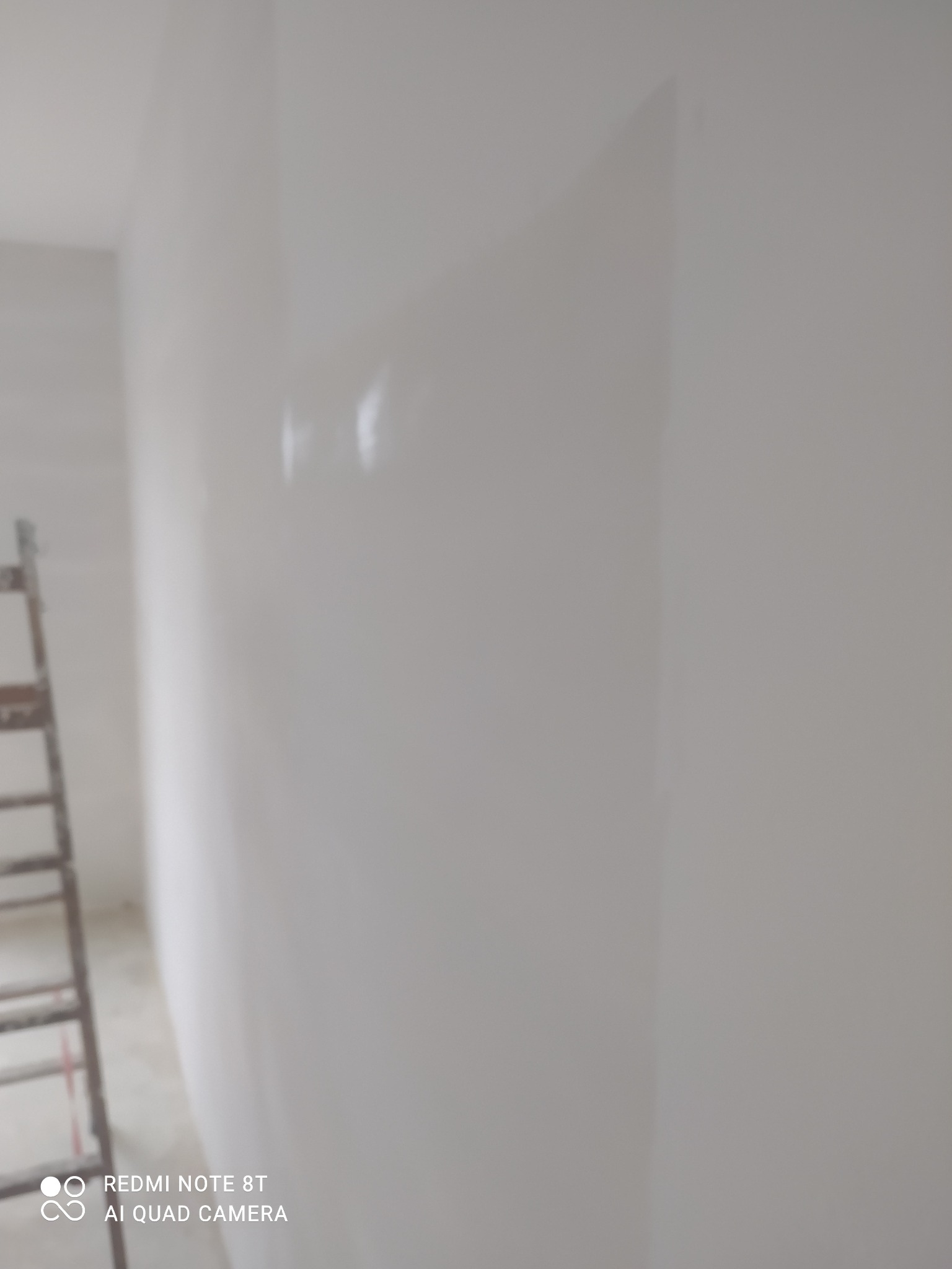 Świeżo pomalowana, biała ściana z delikatnymi refleksami światła, widoczna drabina malarska w lewym dolnym rogu, sugerująca prace malarskie w trakcie realizacji.