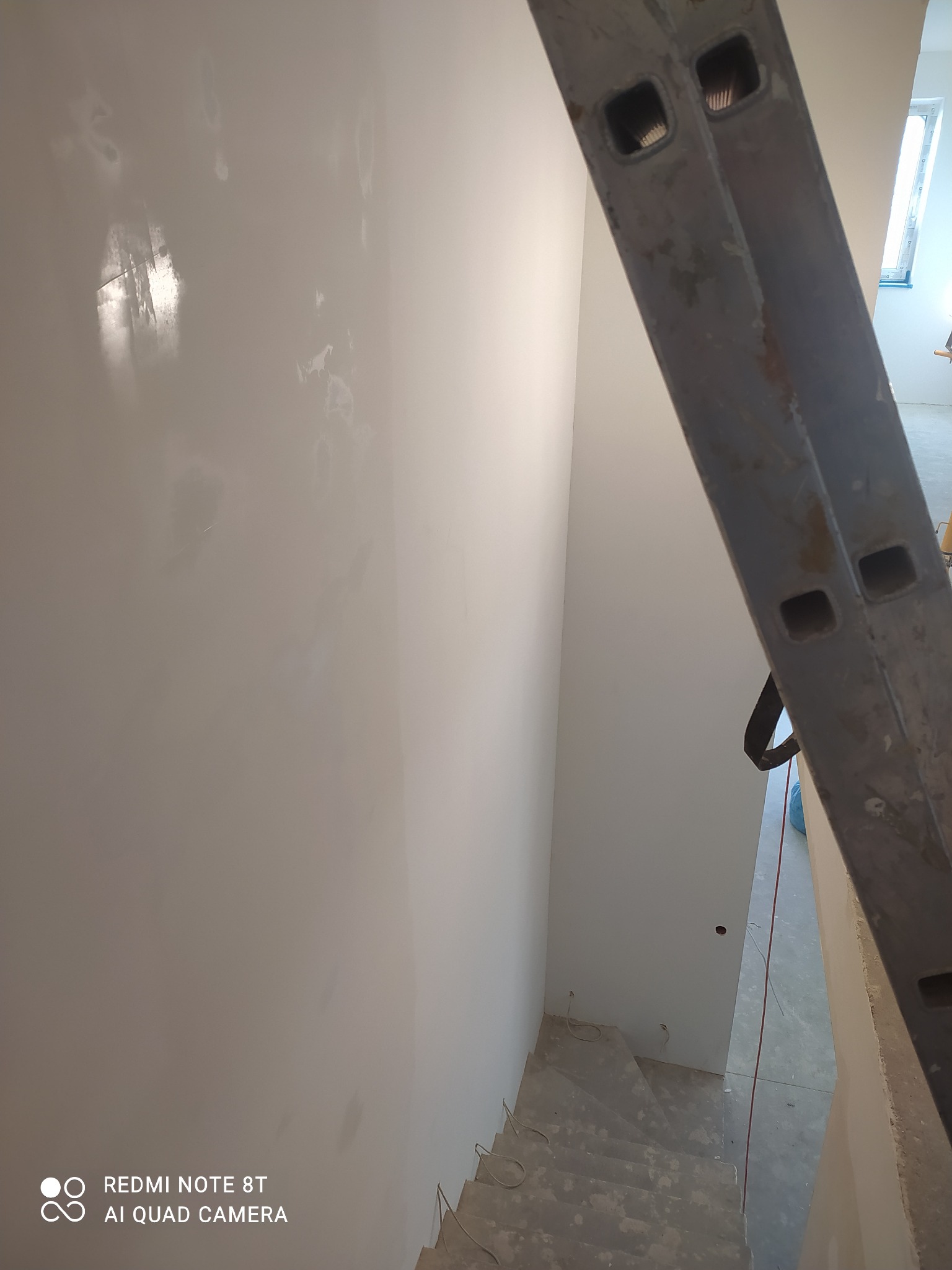 Ściana po szpachlowaniu z widocznymi śladami po łataniu, drabina malarska w kadrze, widok z góry na schody w tle.