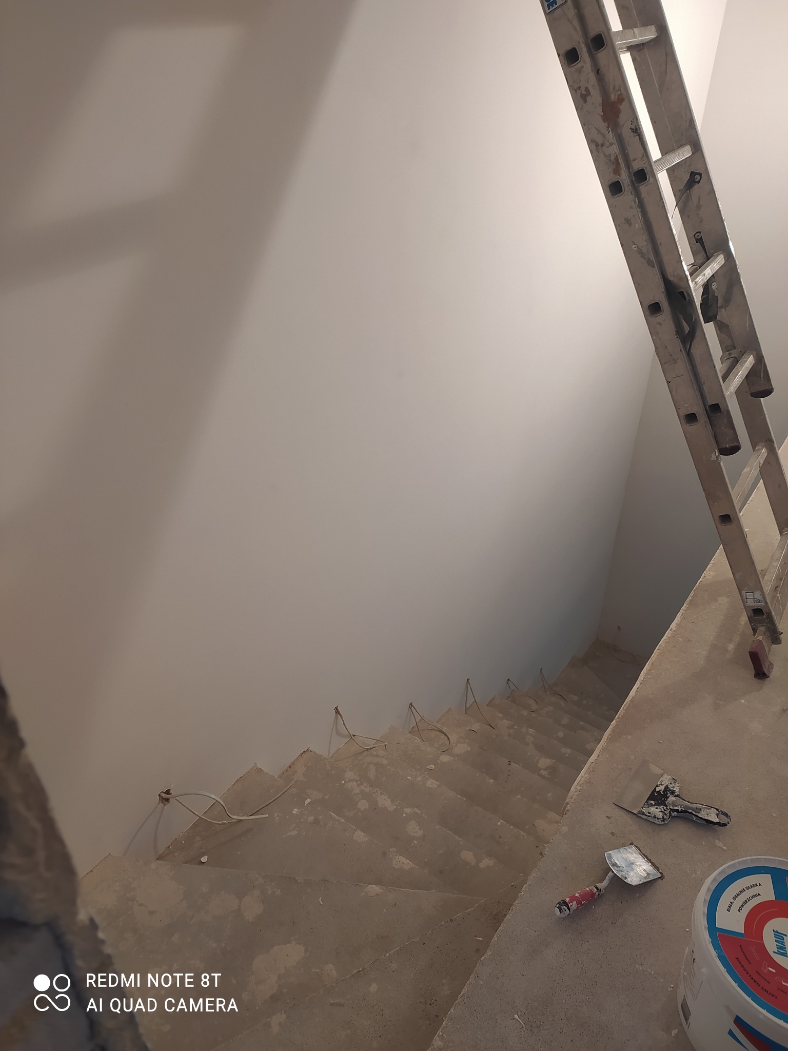 Klatka schodowa w trakcie prac wykończeniowych: świeżo pomalowane ściany, nieukończone schody, drabina aluminiowa oparta o ścianę, widoczne narzędzia szpachlarskie i wiadro z farbą Knauf. Przewody...