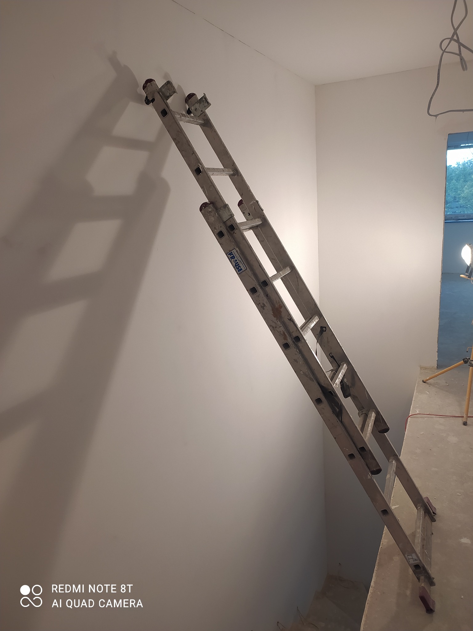 Wnętrze w trakcie prac wykończeniowych, biała ściana z widoczną drabiną aluminiową opartą o nią pod kątem, lampa budowlana na trójnogu w tle.