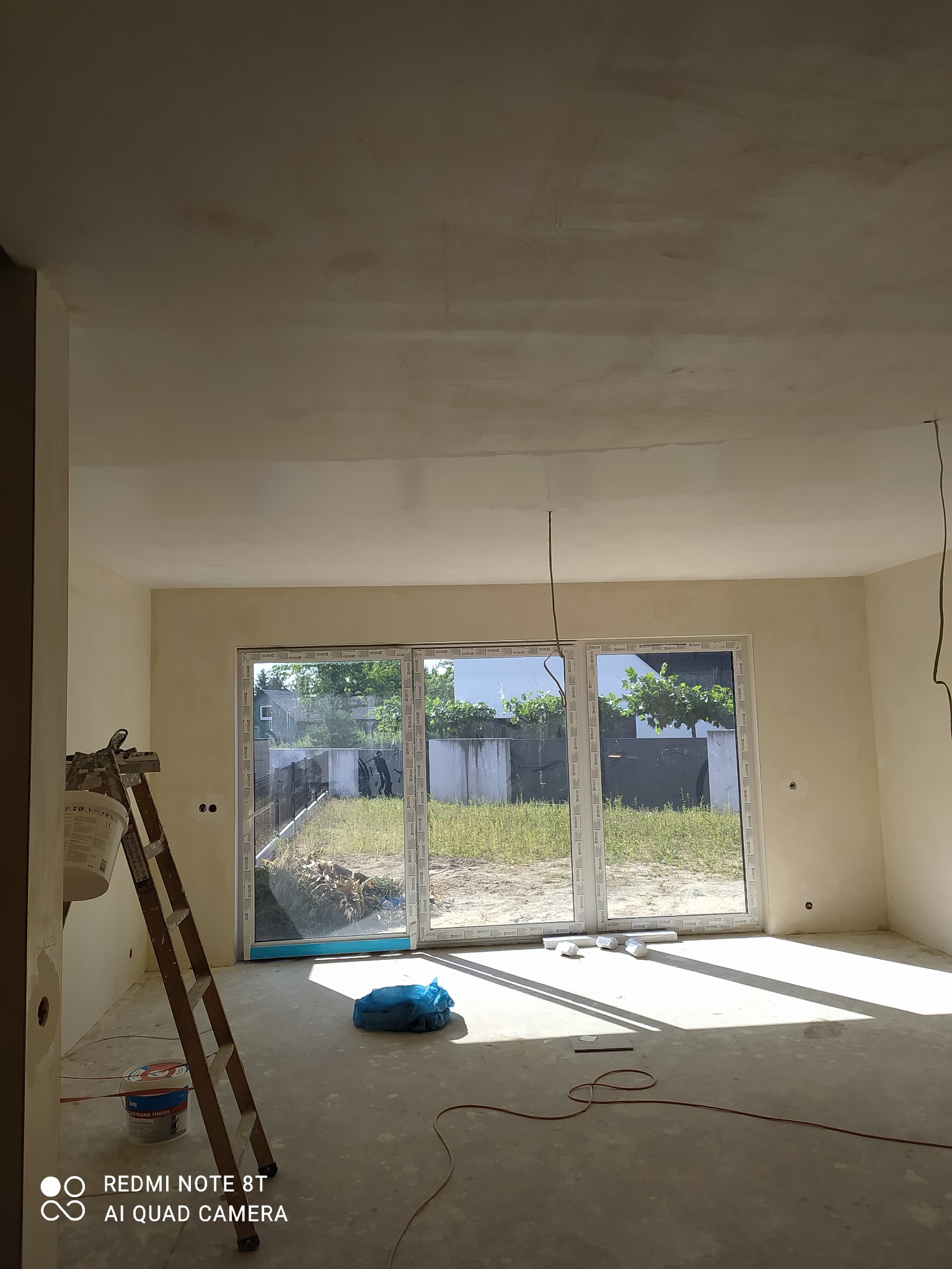Wnętrze w trakcie prac wykończeniowych: szpachlowana ściana i sufit, drabina oparta o ścianę, wiadro z farbą na podłodze, duże okno tarasowe z widokiem na ogród.