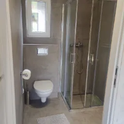 Nowoczesna łazienka z szarą, betonową ścianą i podłogą, kabiną prysznicową ze srebrnymi profilami, białą toaletą i oknem oraz szarym dywanikiem podłogowym.