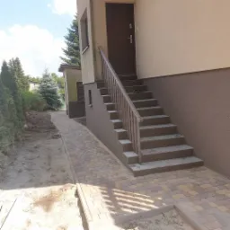 Ułożona kostka brukowa tworząca ścieżkę i schody prowadzące do wejścia domu z brązowymi drzwiami, drewnianą barierką i dwukolorową elewacją.