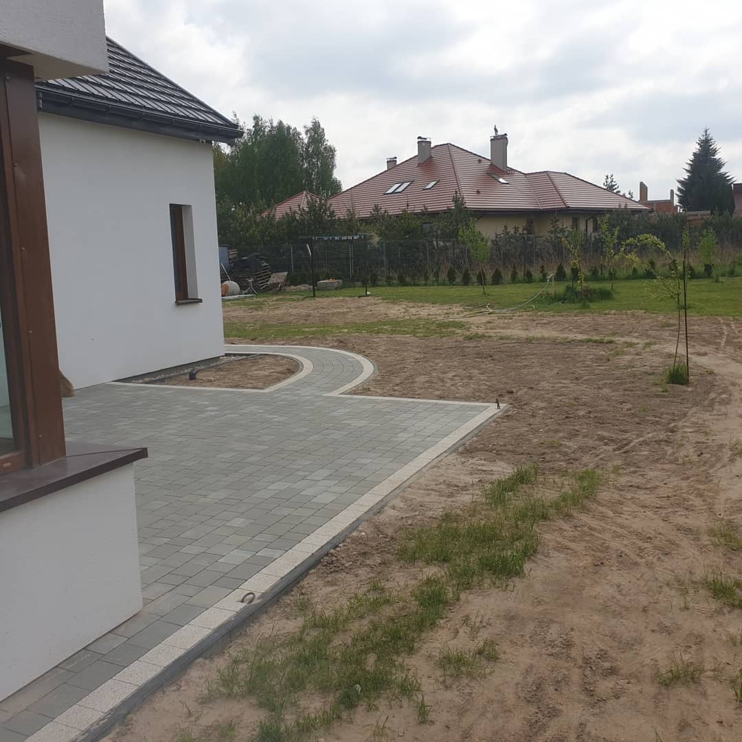 Nowo ułożona, szara kostka brukowa prowadząca od ściany domu w głąb ogrodu, z widocznym przygotowanym terenem pod trawnik.