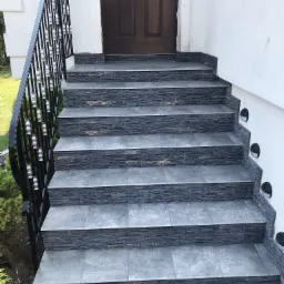 Schody wejściowe wyłożone szarą płytką imitującą kamień, czarna balustrada z ozdobnymi elementami, brązowe drzwi wejściowe, oświetlenie punktowe wbudowane w ścianę obok schodów.