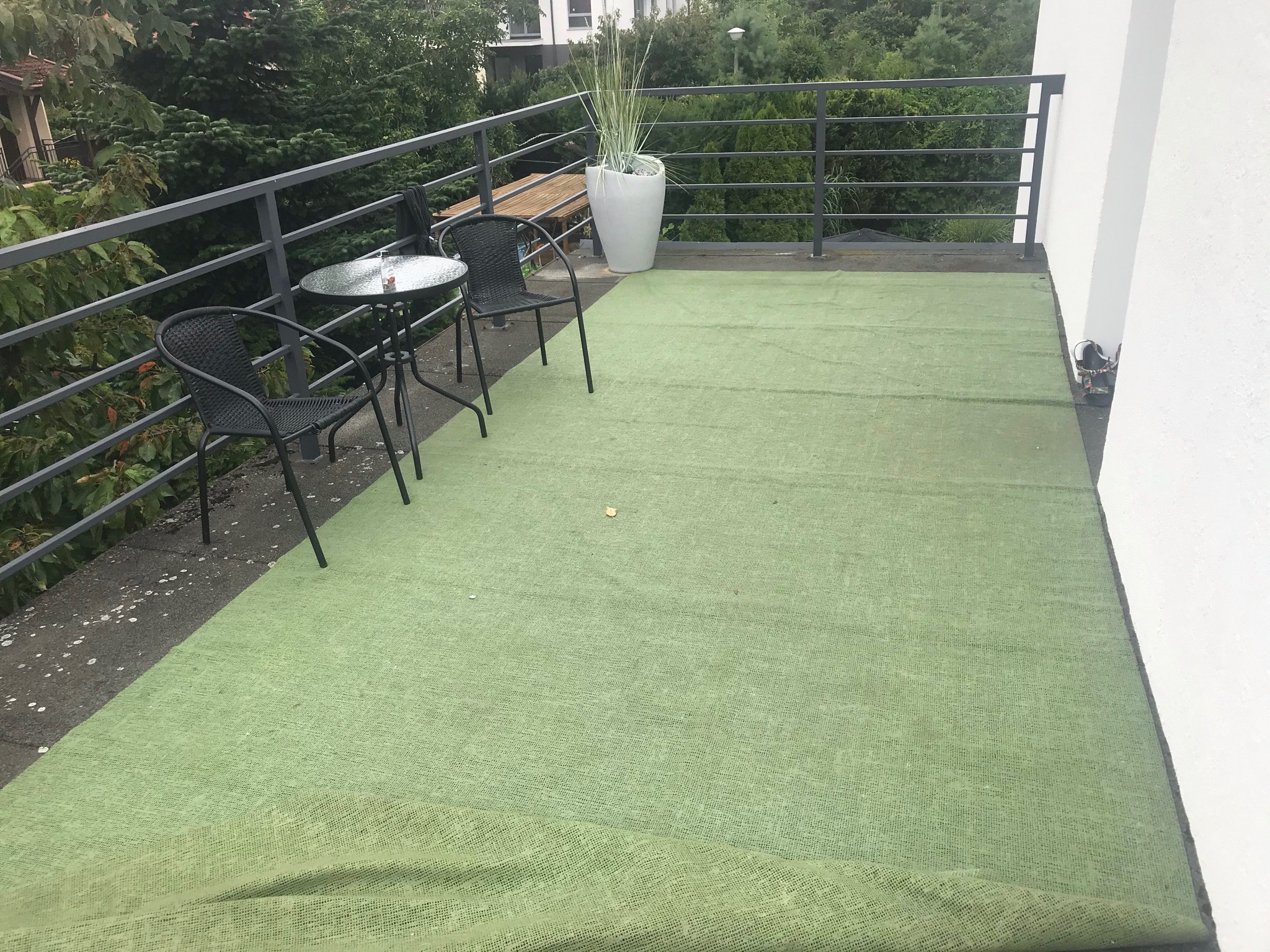 Zadaszony taras z zieloną wykładziną dywanową, czarnymi krzesłami i stolikiem kawowym, białą donicą z wysoką trawą ozdobną oraz metalową balustradą.