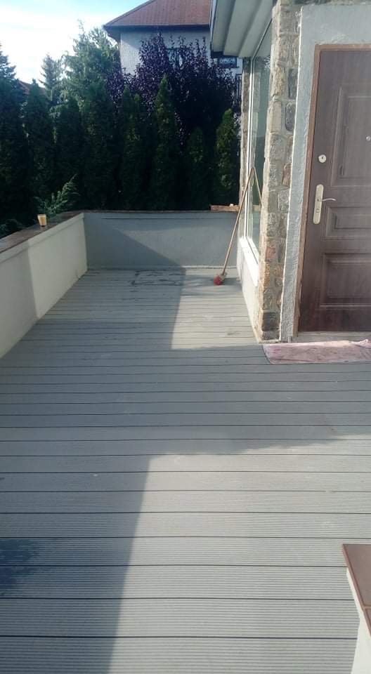 Szara deska kompozytowa na tarasie, widoczna ściana z jasnego betonu, drzwi wejściowe i ceglana elewacja budynku, częściowo zacieniona powierzchnia.