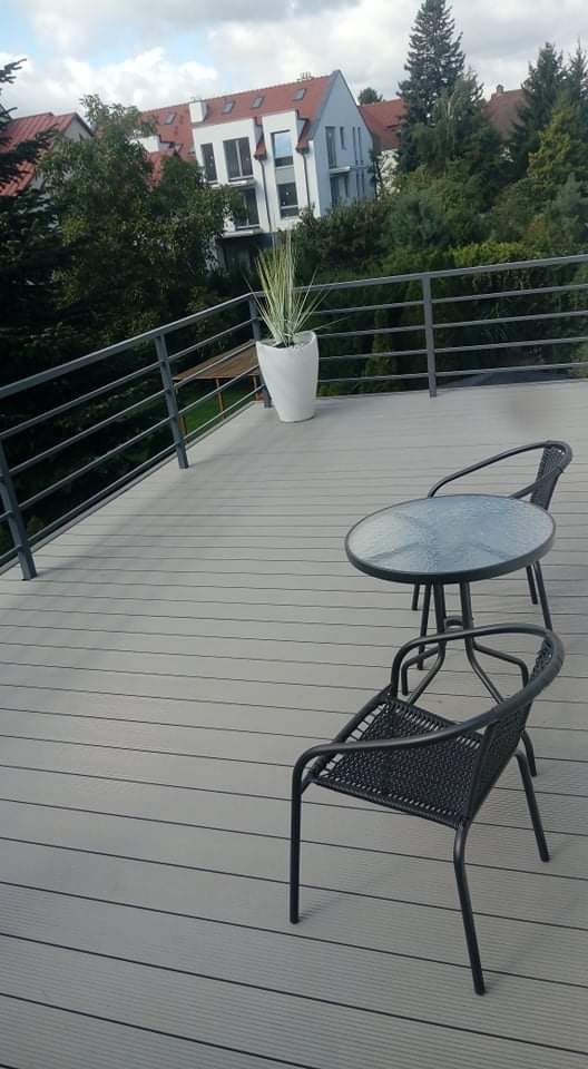 Szary taras z desek kompozytowych z metalową balustradą, okrągły stolik kawowy i dwa krzesła z plecionki, w tle nowoczesny budynek mieszkalny i zieleń.