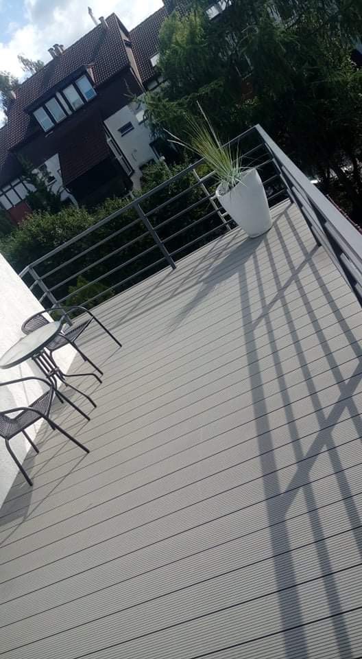 Szary taras z desek kompozytowych, metalowa balustrada, okrągły stolik z dwoma krzesłami i donica z trawą ozdobną, cień rzucany przez balustradę.