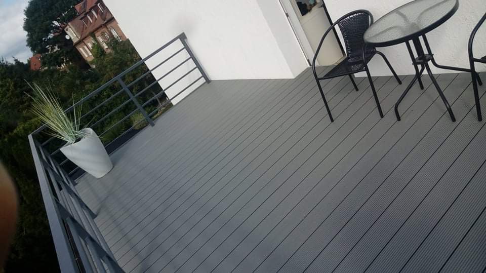 Szary taras kompozytowy z widokiem na zieleń, czarne meble ogrodowe i donica z trawą ozdobną na balkonie.