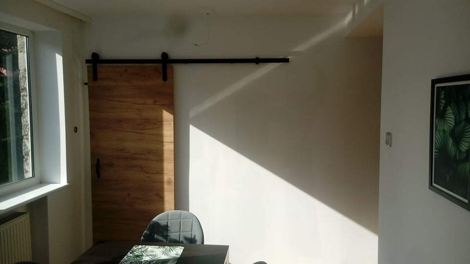 Drewniane drzwi przesuwne na czarnej szynie w jasnym wnętrzu, oświetlone naturalnym światłem, widoczny fragment szarego krzesła i obraz z motywem roślinnym na ścianie.