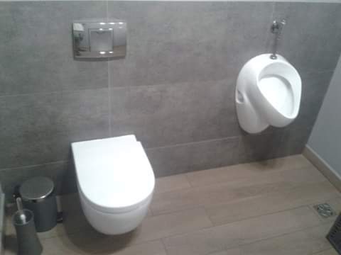 Nowoczesna łazienka z szarymi płytkami, wiszącą toaletą, pisuar i drewnopodobną podłogą, widoczny kosz na śmieci i szczotka do WC.