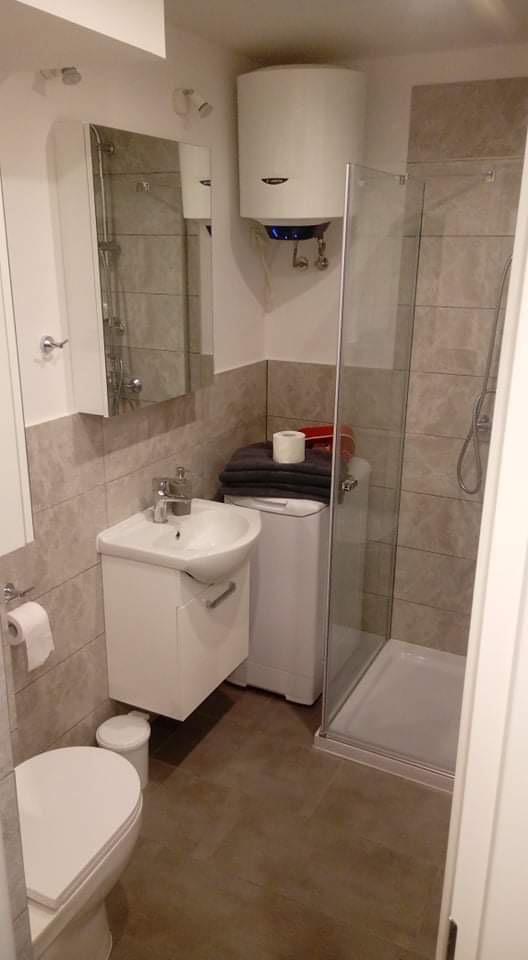 Jasna łazienka z kabiną prysznicową, umywalką z szafką, toaletą, pralką, bojlerem i szarą glazurą w dwóch odcieniach.