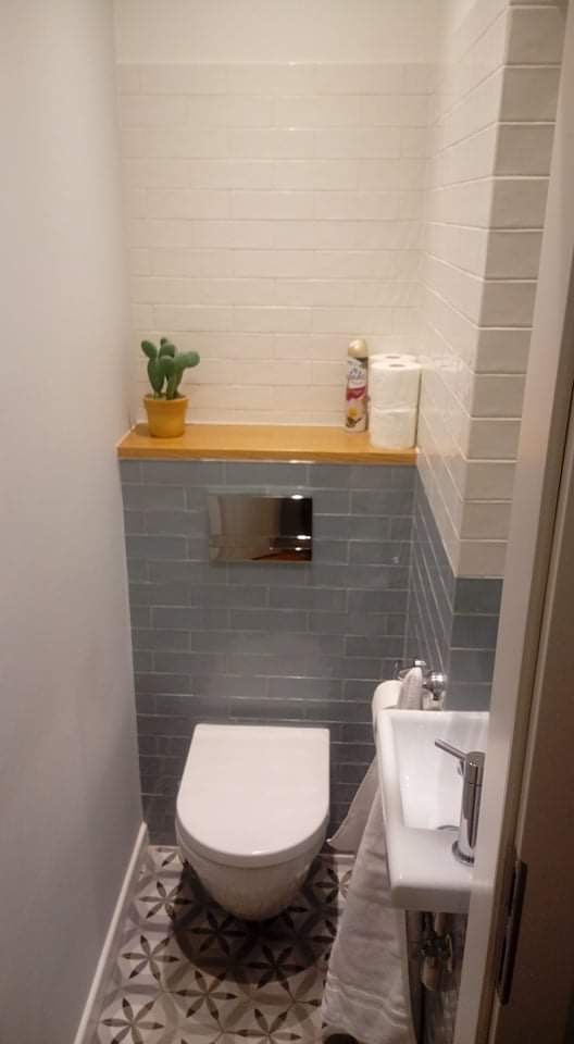 Kompaktowa toaleta z podtynkowym systemem spłukiwania, wykończona białymi i szarymi płytkami imitującymi cegłę, z dekoracyjną podłogą w geometryczne wzory i małą umywalką narożną, całość dopełniona...