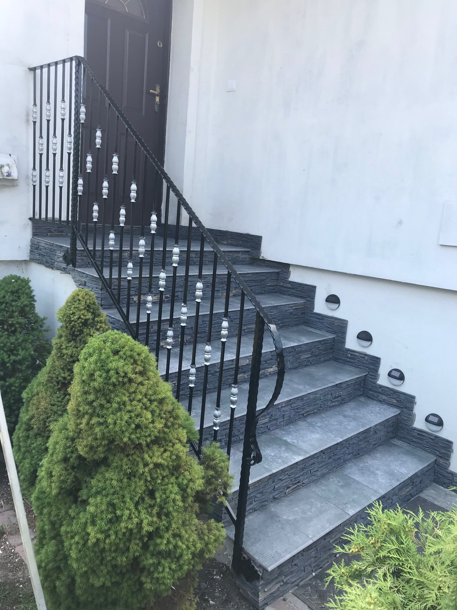 Zewnętrzne schody wejściowe wyłożone szarą płytką imitującą kamień, z czarną metalową balustradą z ozdobnymi elementami i oświetleniem punktowym w ścianie, otoczone zielonymi krzewami.