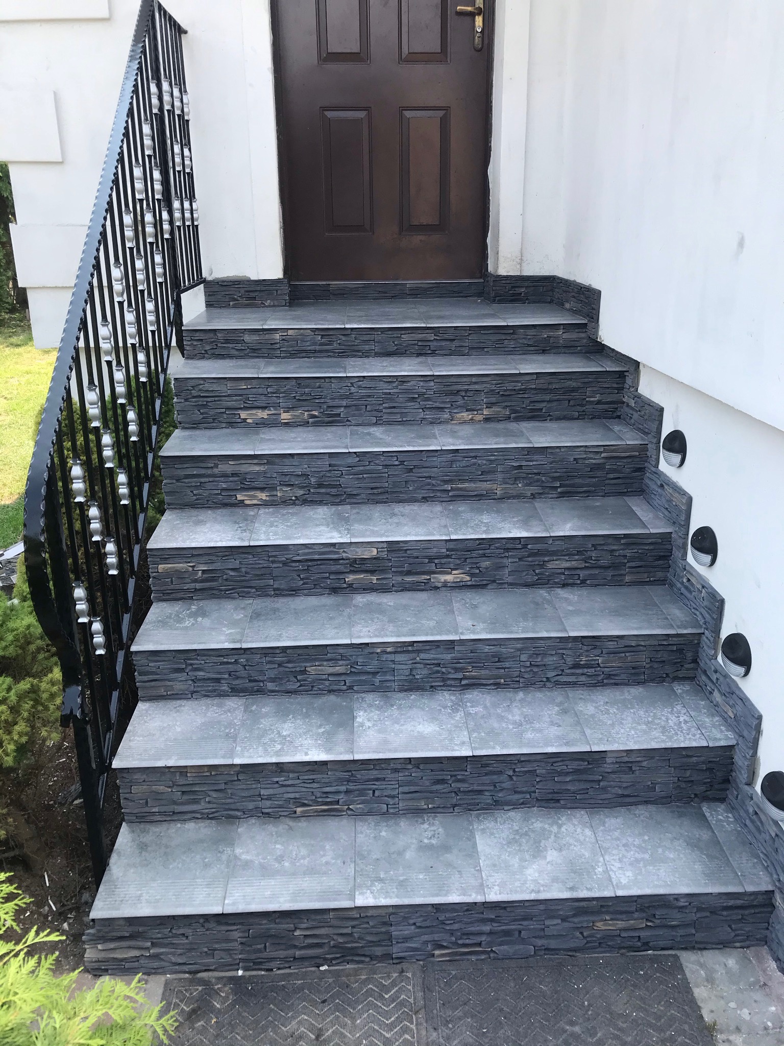 Schody wejściowe wyłożone szarą płytką imitującą kamień, czarna balustrada z ozdobnymi elementami, brązowe drzwi wejściowe, oświetlenie punktowe wbudowane w ścianę obok schodów.