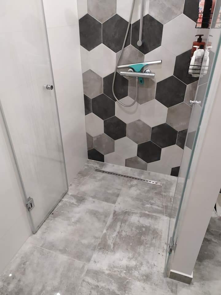 Wykończona kabina prysznicowa z heksagonalnymi płytkami w odcieniach szarości i bieli na ścianie, szare płytki podłogowe imitujące beton, odpływ liniowy i szklane drzwi.